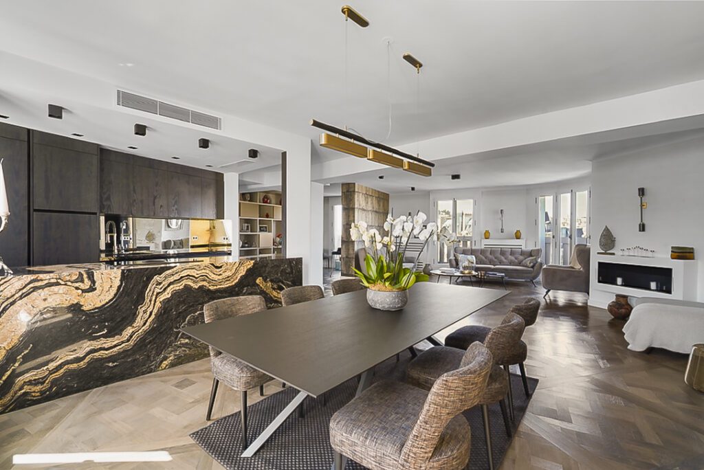 Appartement luxueux à Paris avec cuisine moderne en marbre, comptoir élégant et équipements haut de gamme. Une grande table à manger est entourée de chaises en rotin, et un arrangement floral ajoute une touche de sophistication. L'espace ouvert offre une vue dégagée et une décoration soignée.