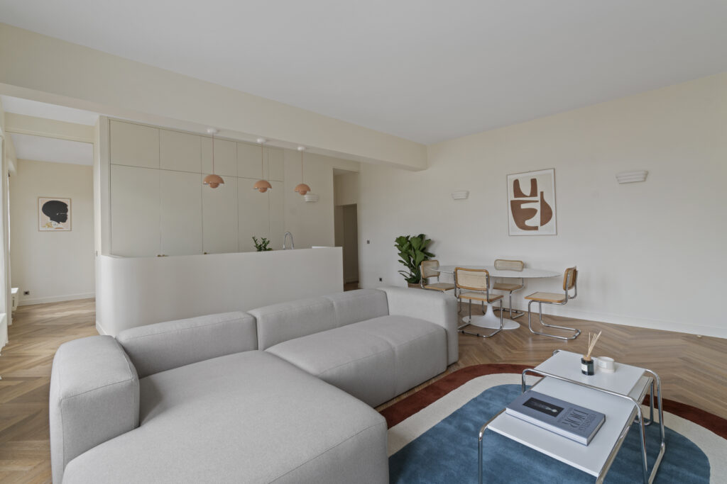 Salon lumineux d'une maison luxueuse avec de grandes fenêtres donnant sur un jardin. L'espace est décoré avec un canapé élégant, des fauteuils au motif léopard et un tapis oriental. Les colonnes et les rideaux gris ajoutent une touche sophistiquée à la pièce.