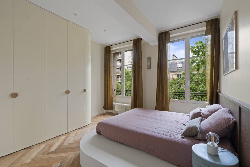 Chambre moderne à Paris avec parquet en chevron, lit avec draps violets et meubles aux tons neutres. Les grandes fenêtres laissent entrer la lumière naturelle, offrant une vue sur la ville. Le design épuré et les rideaux dorés créent une atmosphère chaleureuse.