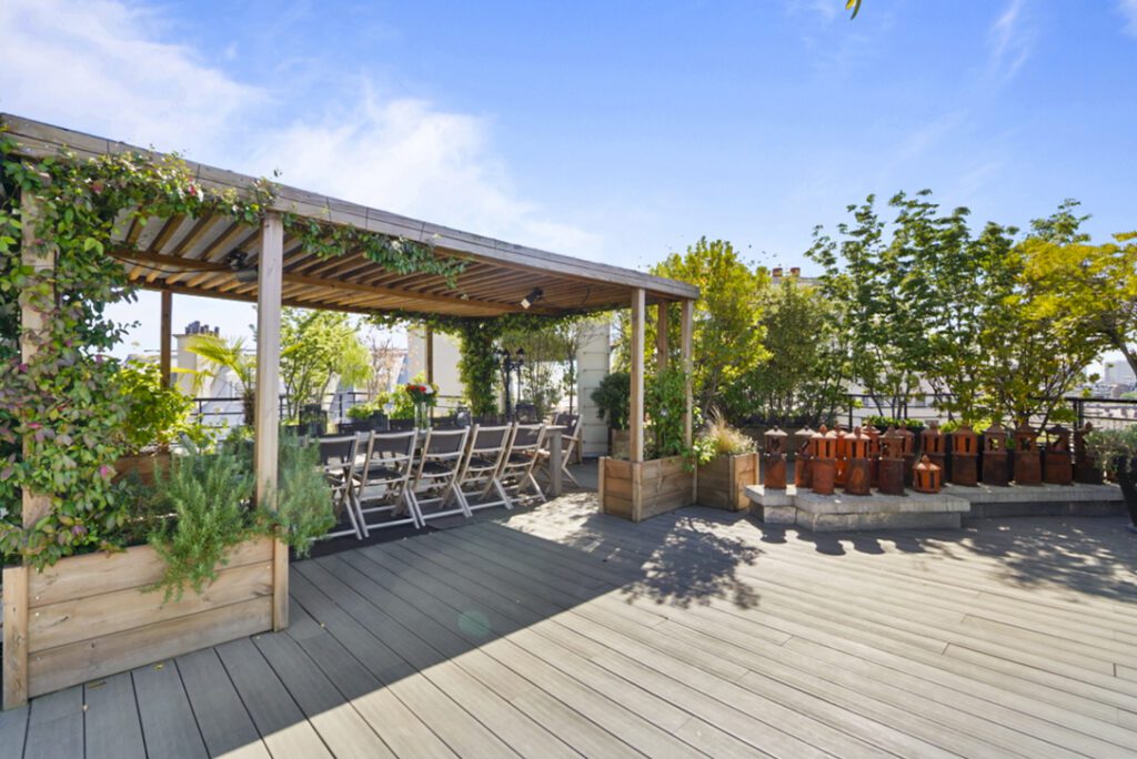 Terrasse végétalisée à Paris, avec un pergola en bois abritant une grande table en extérieur et des chaises pliantes. Des plantes luxuriantes et des pots de fleurs ajoutent une touche de verdure à l'espace, offrant une vue dégagée sur la ville.