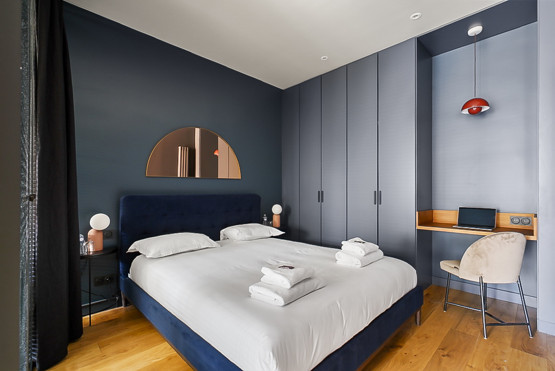 Chambre moderne dans un appartement Airbnb à Bordeaux, avec un lit king-size bleu, un miroir rond au-dessus du lit et des rangements intégrés. Une lampe suspendue au plafond et un petit bureau avec un ordinateur portable ajoutent une touche fonctionnelle et élégante à l'espace.