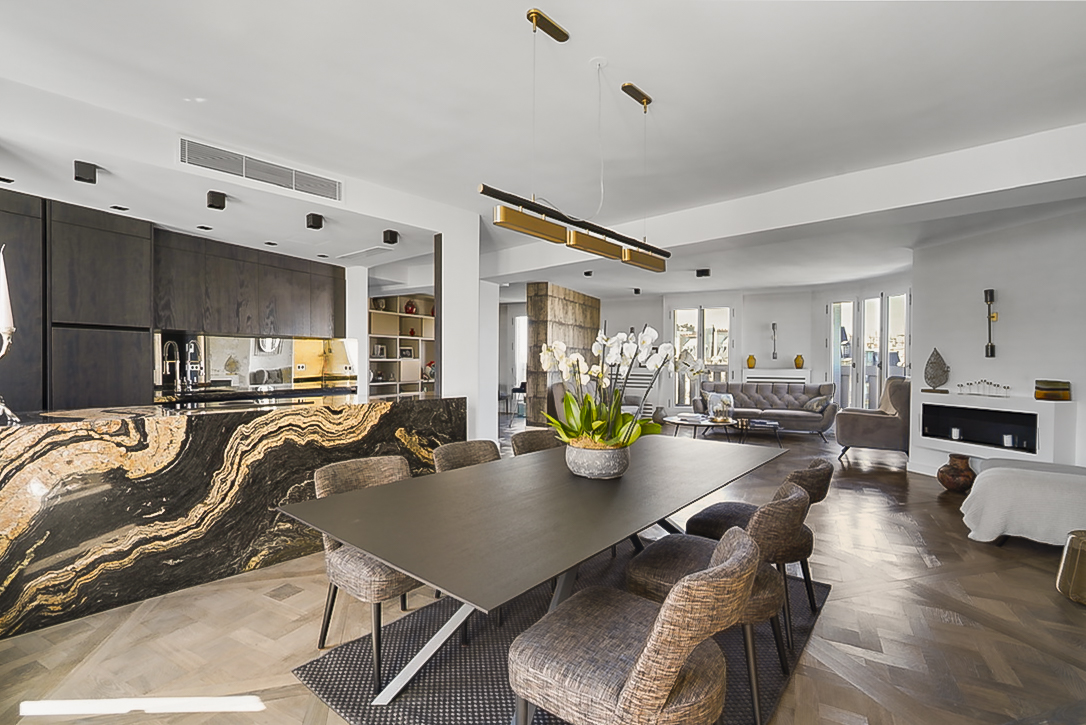 Appartement luxueux à Paris avec cuisine moderne en marbre, comptoir élégant et équipements haut de gamme. Une grande table à manger est entourée de chaises en rotin, et un arrangement floral ajoute une touche de sophistication. L'espace ouvert offre une vue dégagée et une décoration soignée.