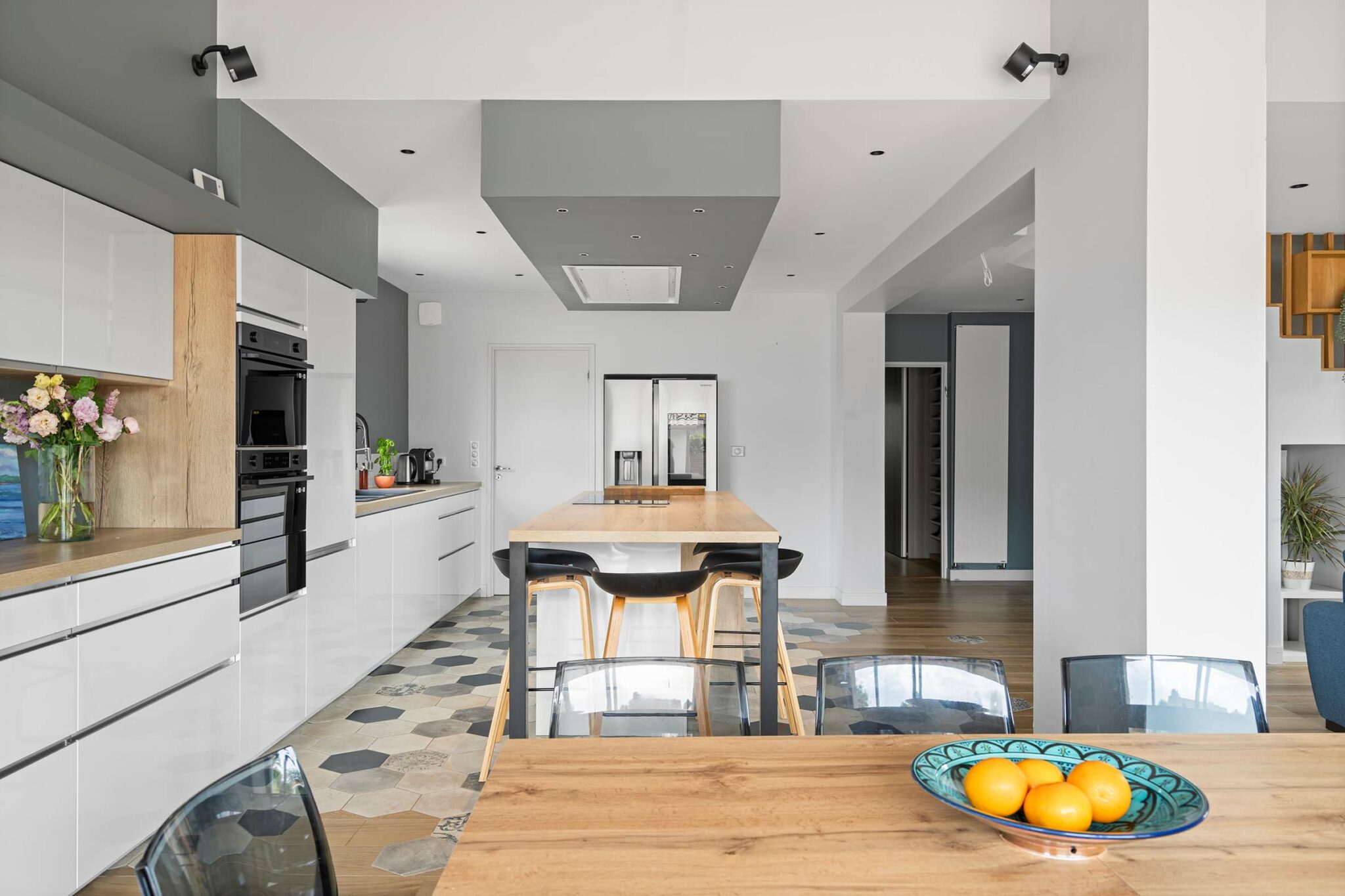 Cuisine moderne avec îlot central en bois et chaises noires. Le sol est décoré de carreaux hexagonaux, et un bol de fruits est posé sur la table en bois. L'éclairage intégré au plafond ajoute une touche moderne à l'espace.