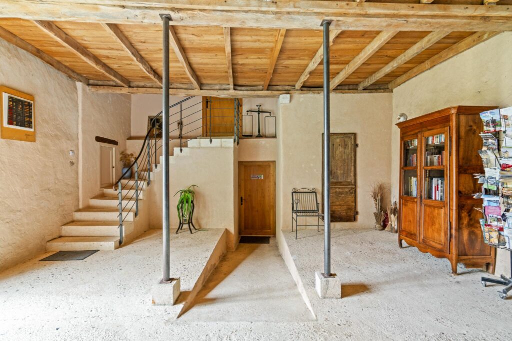 Entrée d'un gîte avec un escalier en pierre menant à l'étage. Le sol est en béton brut, et les poutres en bois du plafond ajoutent une touche rustique à l'espace. Une bibliothèque en bois et une chaise métallique décorent la pièce, créant une atmosphère chaleureuse et accueillante.