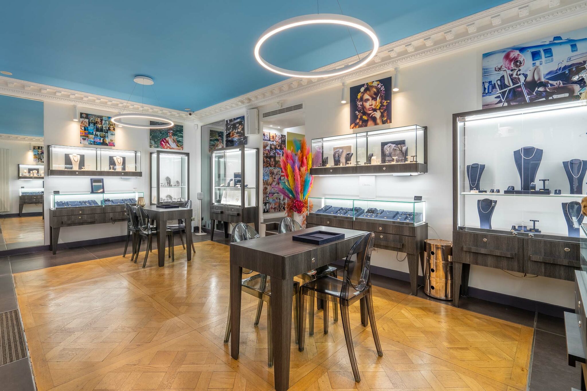 Boutique de bijoux moderne à Paris, avec des vitrines lumineuses présentant des pièces de luxe. L'intérieur est élégant, avec un parquet en chevron, des meubles en bois foncé et des chaises transparentes. L'éclairage circulaire au plafond ajoute une touche professionnelle à l'espace.