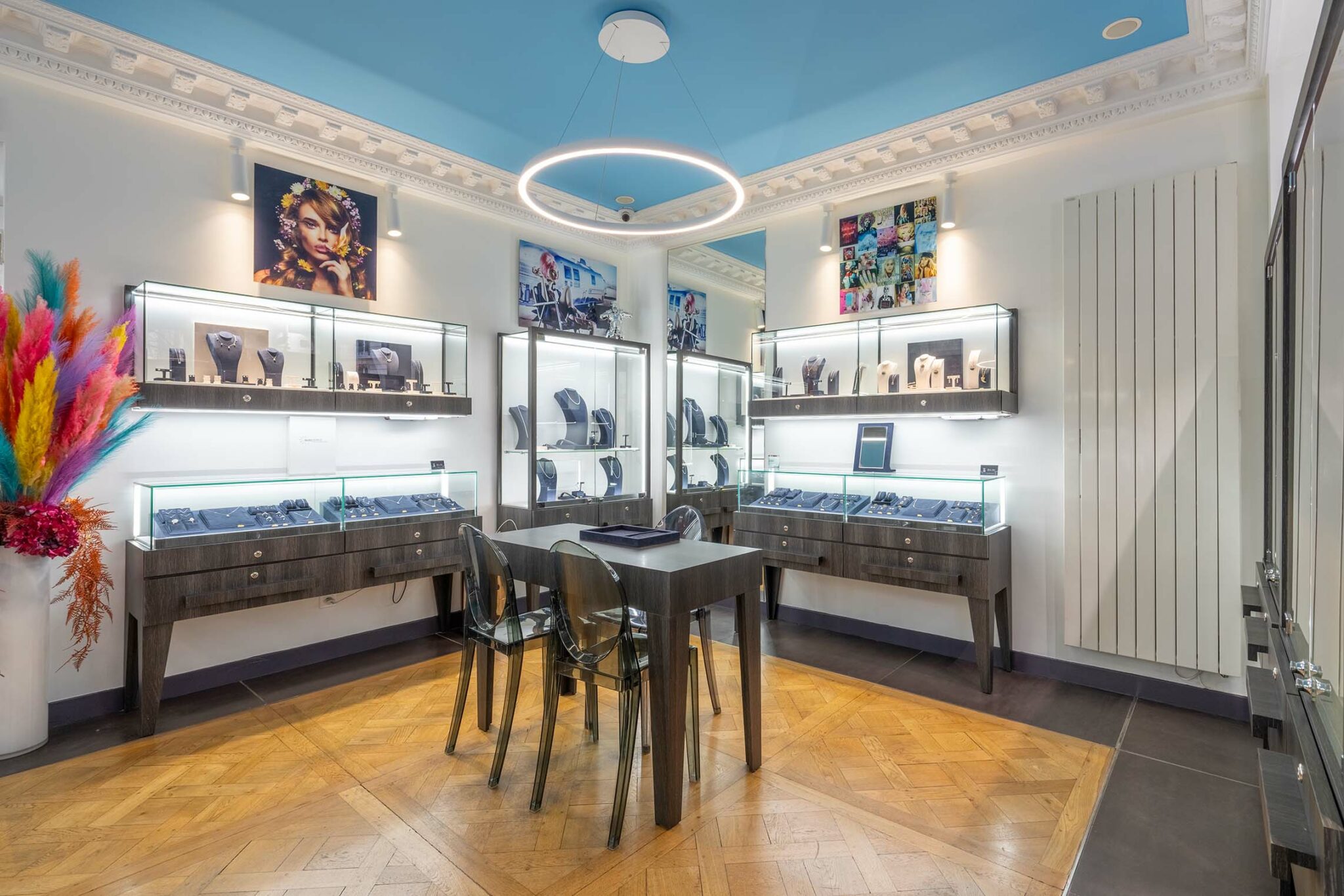 Boutique de bijoux moderne à Paris, avec des vitrines lumineuses présentant des pièces de luxe. L'intérieur est élégant, avec un parquet en chevron, des meubles en bois foncé et des chaises transparentes. L'éclairage circulaire au plafond ajoute une touche professionnelle à l'espace.