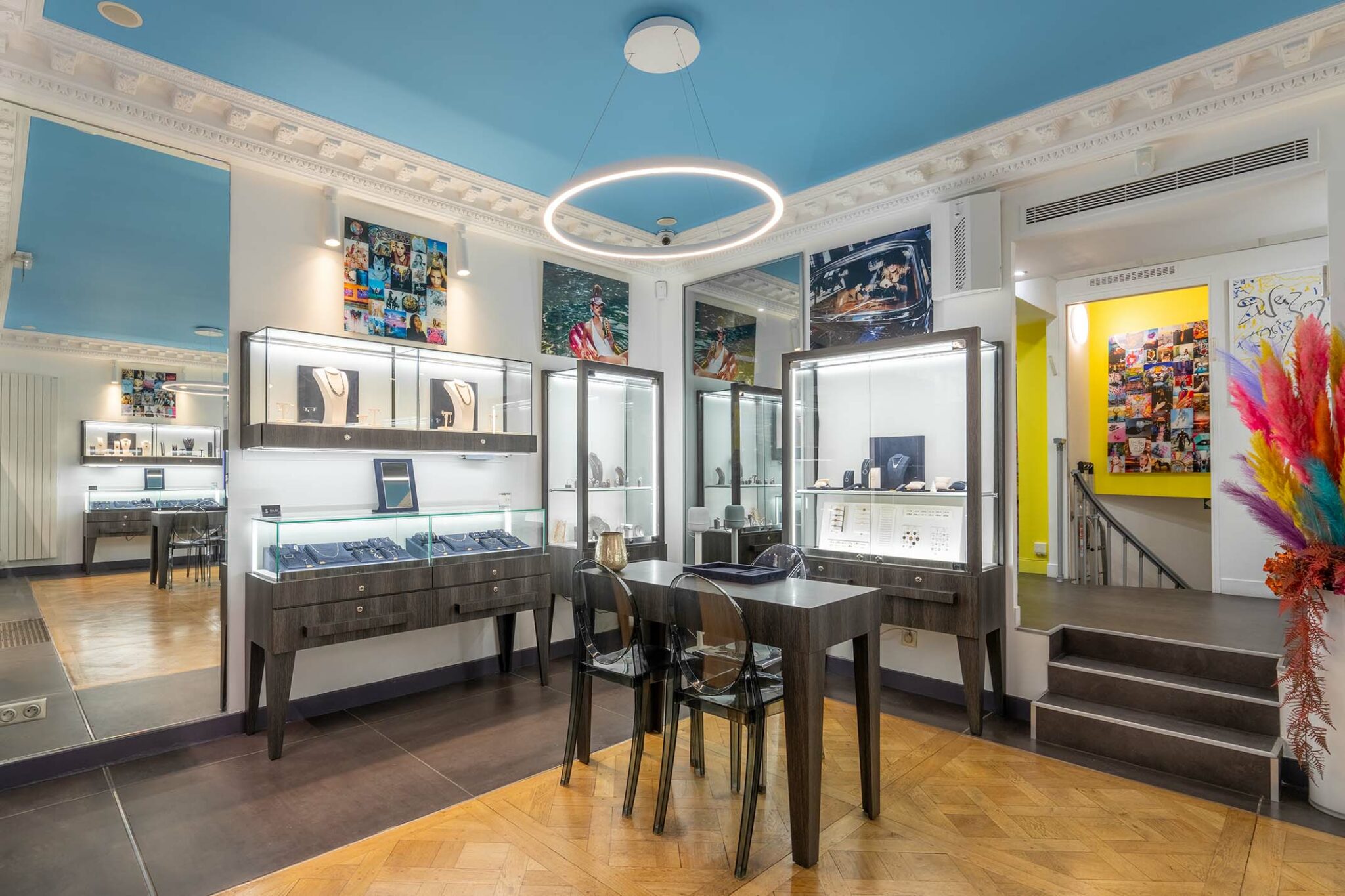Boutique de bijoux moderne à Paris, avec des vitrines lumineuses présentant des pièces de luxe. L'intérieur est élégant, avec un parquet en chevron, des meubles en bois foncé et des chaises transparentes. L'éclairage circulaire au plafond ajoute une touche professionnelle à l'espace.