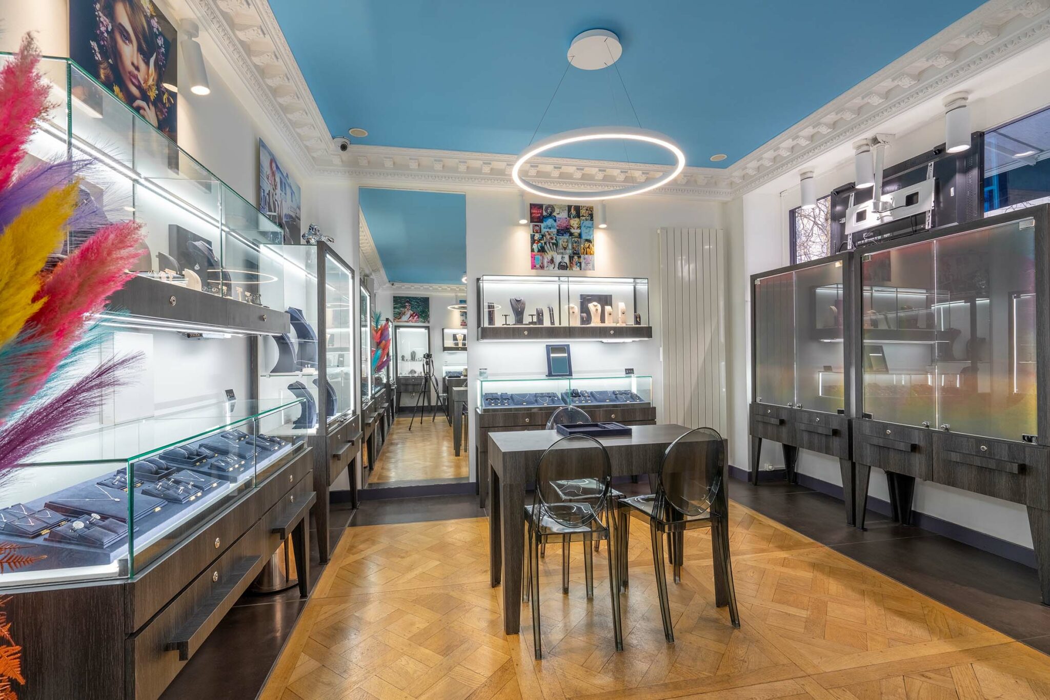 Boutique de bijoux moderne à Paris, avec des vitrines lumineuses présentant des pièces de luxe. L'intérieur est élégant, avec un parquet en chevron, des meubles en bois foncé et des chaises transparentes. L'éclairage circulaire au plafond ajoute une touche professionnelle à l'espace.